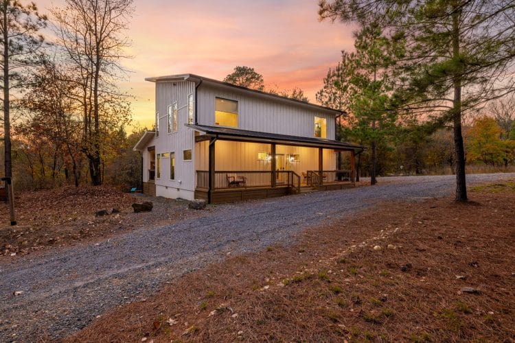 Front of the white two-story cabin with a metal roof and long porch, gravel drive curving past at sunset
