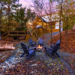 Fire pit with dark Adirondack chairs at dusk, a small wooden footbridge and the deck stairs behind