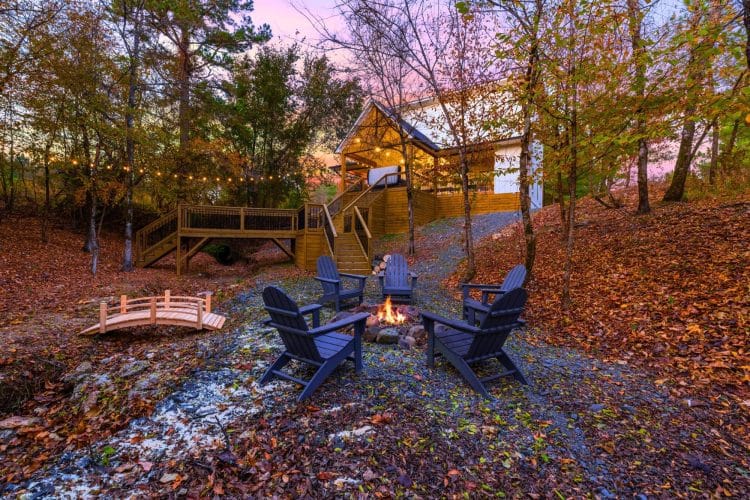 Fire pit with dark Adirondack chairs at dusk, a small wooden footbridge and the deck stairs behind