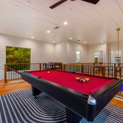 Pool table with red felt in the upstairs loft, with a ceiling fan, railing and a window behind it