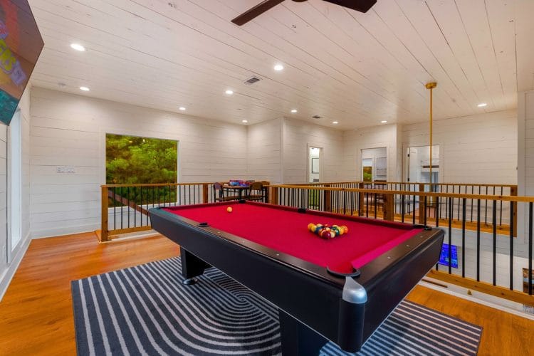 Pool table with red felt in the upstairs loft, with a ceiling fan, railing and a window behind it