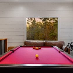 Red-felt pool table with a window, a leather loveseat and cue sticks on the wall