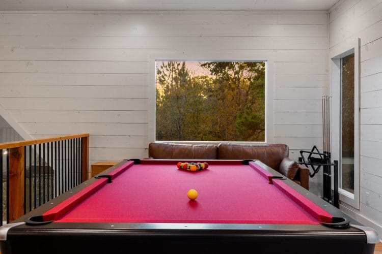Red-felt pool table with a window, a leather loveseat and cue sticks on the wall