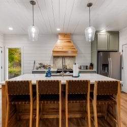 Kitchen island with four woven bar stools, sage-green cabinets, two glass pendant lights and a wood range hood