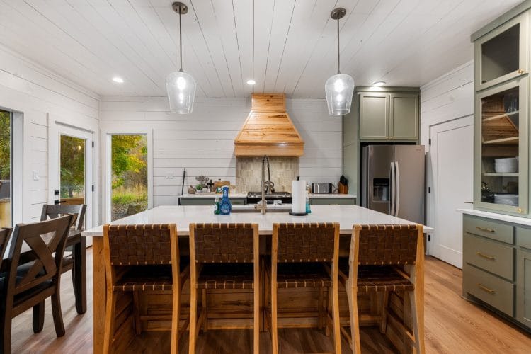 Kitchen island with four woven bar stools, sage-green cabinets, two glass pendant lights and a wood range hood