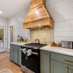 Range under a wood hood, with sage-green cabinets, white quartz counters and a door to the deck