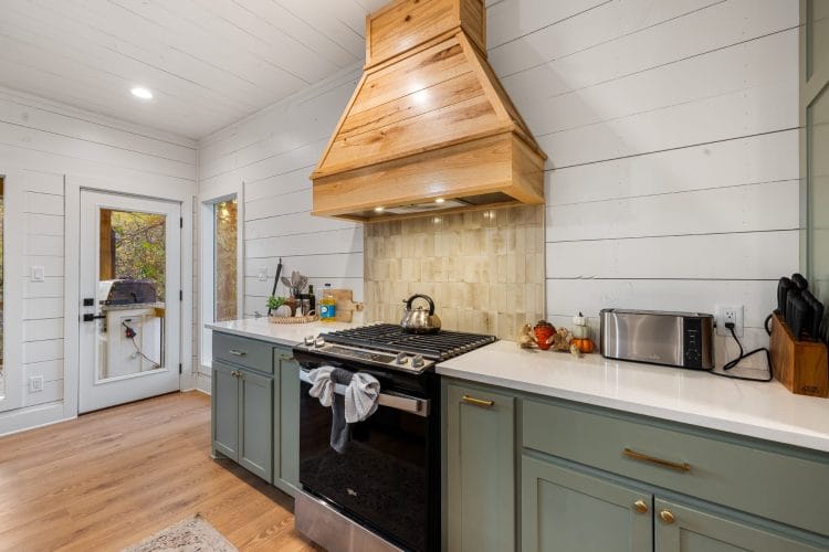Range under a wood hood, with sage-green cabinets, white quartz counters and a door to the deck