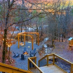 Gravel yard seen from the deck, with a fire pit ringed by Adirondack chairs, a swing frame, a playset and a small creek bed under string lights