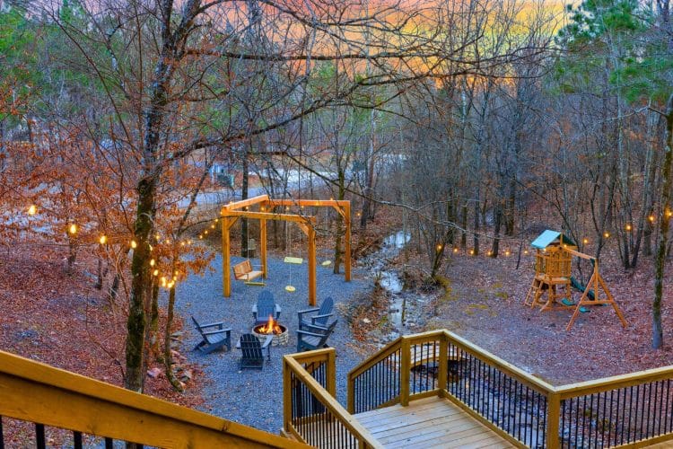 Gravel yard seen from the deck, with a fire pit ringed by Adirondack chairs, a swing frame, a playset and a small creek bed under string lights