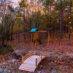 Playset with a slide beyond a small wooden bridge over a creek bed, with string lights in the trees
