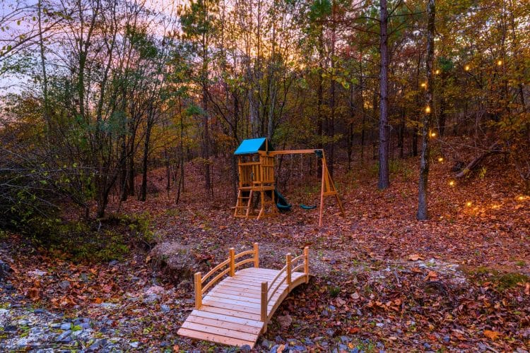 Playset with a slide beyond a small wooden bridge over a creek bed, with string lights in the trees