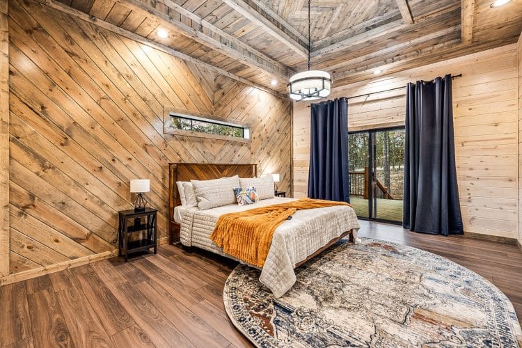King bed with an orange throw and navy curtains beside a sliding glass door, pine walls