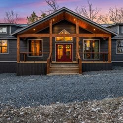 Front of the dark gray single-story cabin with a gabled porch, red front doors and a gravel drive at sunset