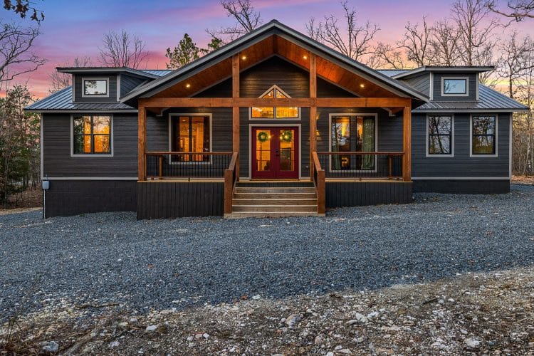 Front of the dark gray single-story cabin with a gabled porch, red front doors and a gravel drive at sunset