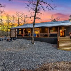 Back of the cabin with a long covered porch, wooden steps and a gravel yard at sunset