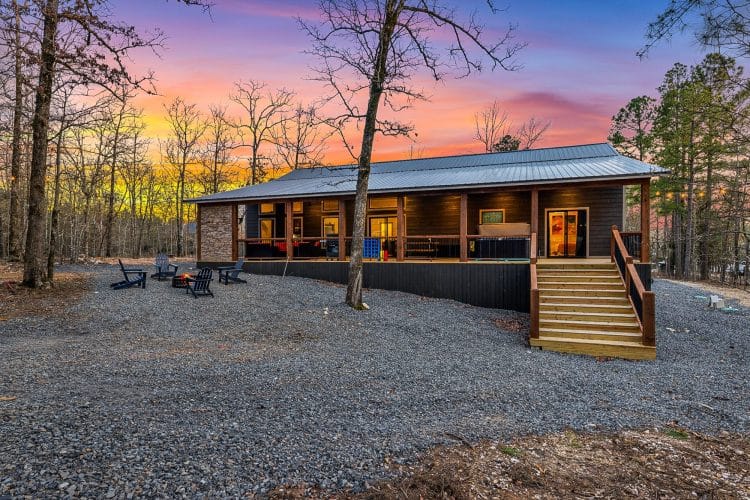 Back of the cabin with a long covered porch, wooden steps and a gravel yard at sunset
