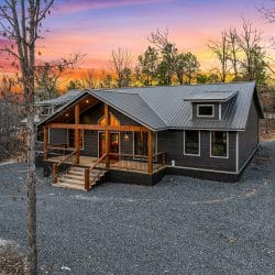 Front of the gray cabin with a gabled porch and red doors seen from the drive at sunset