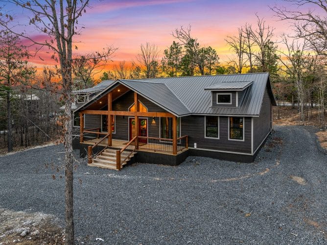 Front of the gray cabin with a gabled porch and red doors seen from the drive at sunset