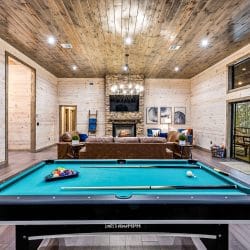Pool table with teal felt in the main room, with a stone fireplace and a pine plank ceiling behind