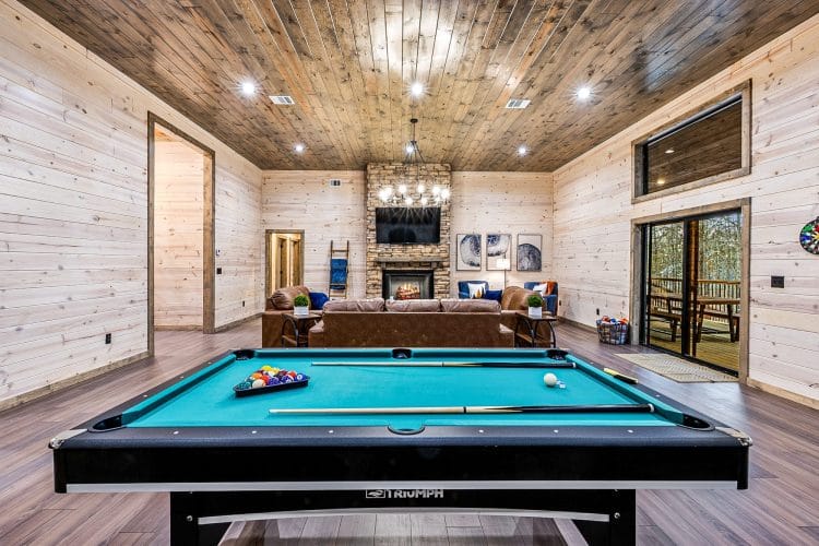 Pool table with teal felt in the main room, with a stone fireplace and a pine plank ceiling behind