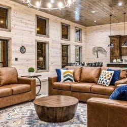 Living room with brown leather sofas, a round wood coffee table and a chandelier, the kitchen behind