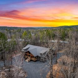 Aerial of the cabin and gravel drive among bare trees at sunset