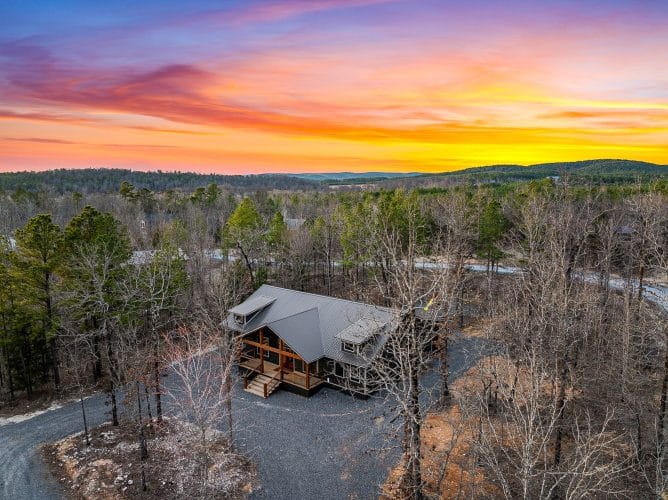 Aerial of the cabin and gravel drive among bare trees at sunset