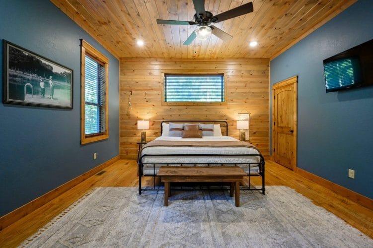 King bed with a dark metal frame and wood bench on a gray rug, TV on the right wall