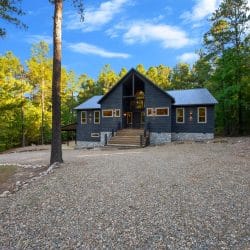 Charcoal-gray cabin with a stone base and front steps at the end of a gravel drive, tall pines around it