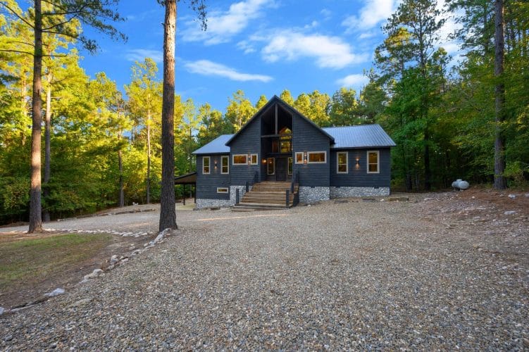 Charcoal-gray cabin with a stone base and front steps at the end of a gravel drive, tall pines around it