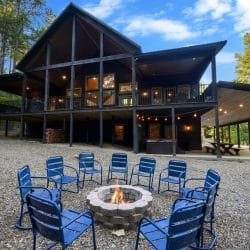 Back of the cabin with the long upper deck, a hot tub under it, a covered picnic area and a fire pit ringed with blue chairs