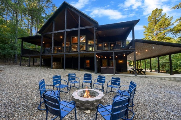 Back of the cabin with the long upper deck, a hot tub under it, a covered picnic area and a fire pit ringed with blue chairs