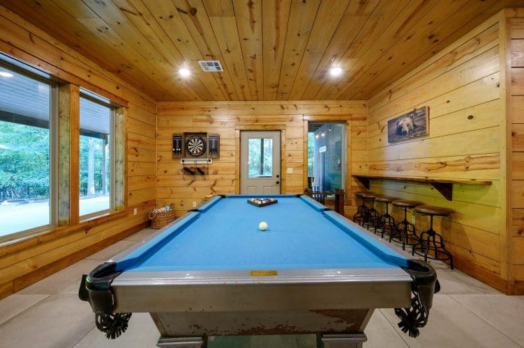 Blue-felt pool table with a dartboard and a row of bar stools along the wall