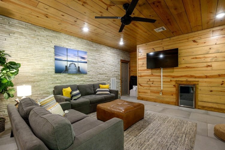 Lower-level living area with gray sofas, a leather ottoman and a wall-mounted TV