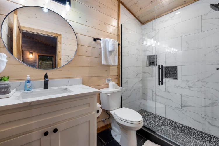 Bathroom with a round mirror, a white vanity and a marble-look shower