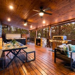 Covered deck with a dining table, a gas grill, a stone fireplace and the hot tub at the far end