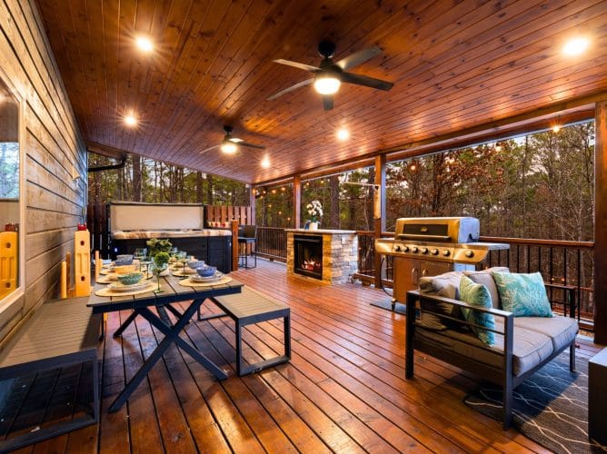Covered deck with a dining table, a gas grill, a stone fireplace and the hot tub at the far end