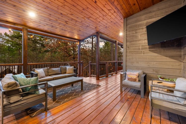 Covered deck with an outdoor sofa and chairs and a big TV on the wall