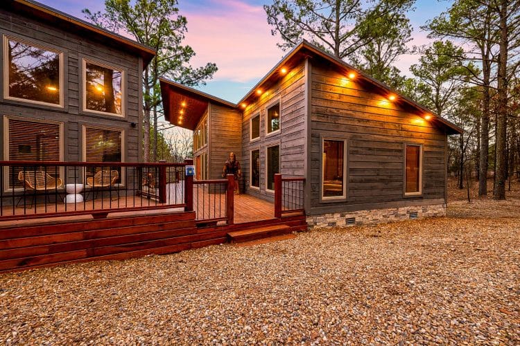 Back of the main house and game house with a large reddish deck and lights at dusk