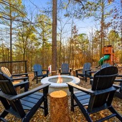 Fire pit with black Adirondack chairs and a stump table, the playset behind in the trees