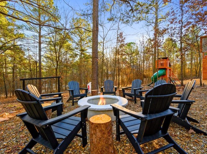 Fire pit with black Adirondack chairs and a stump table, the playset behind in the trees