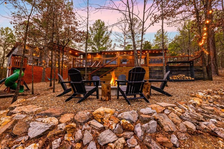 Stone-ringed fire pit with Adirondack chairs and the lit house behind