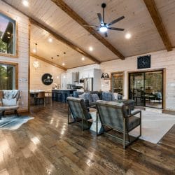 Main house living room with a vaulted pine ceiling, wood beams, gray armchairs and a sliding door to the deck