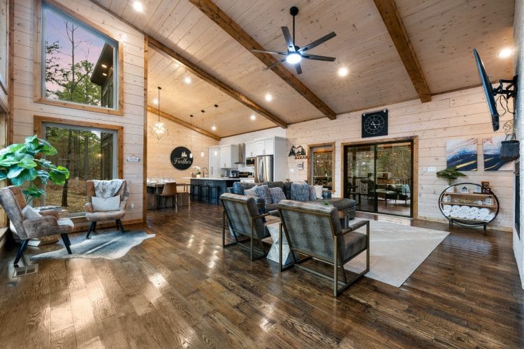 Main house living room with a vaulted pine ceiling, wood beams, gray armchairs and a sliding door to the deck