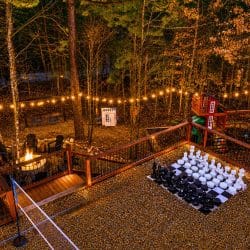 Night view of the back deck and yard with string lights, a fire pit, giant chess on the gravel and a playset
