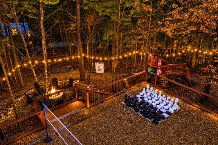 Night view of the back deck and yard with string lights, a fire pit, giant chess on the gravel and a playset