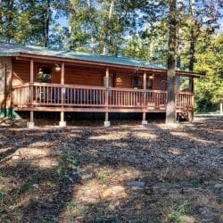 Front of the cabin with a green roof, railed porch and gravel yard under tall trees