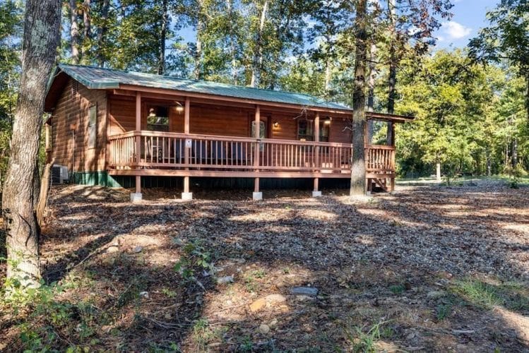 Front of the cabin with a green roof, railed porch and gravel yard under tall trees