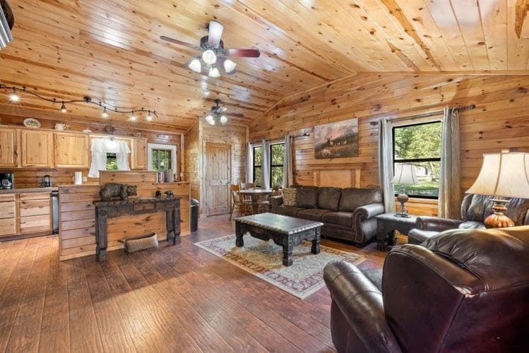 Open living area and kitchen under a pine ceiling with two ceiling fans, a leather sofa and recliners
