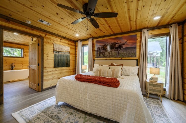 King bed with white quilted bedding under a pine ceiling, a longhorn print and an open bathroom door
